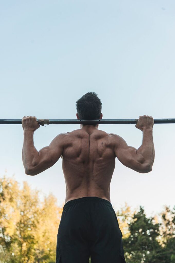Rear view of a strong man exercising shirtless with a barbell outdoors, showcasing strength and fitness.